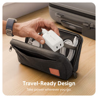 A dark gray zippered travel pouch on a wooden surface, shown open with a hand placing a white power adapter inside. The pouch contains cables in elastic loops, with a gray suitcase in the background and the text 'Travel-Ready Design' at the bottom.