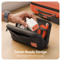 A hand inserts a white and orange power adapter into a dark gray zippered travel pouch containing an orange cable, with an orange suitcase in the background on a wooden surface. Text at the bottom reads 'Travel-Ready Design' and 'Take power wherever you go.