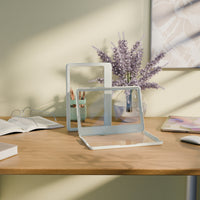 Dos soportes transparentes sobre una superficie de madera, con un jarrón de flores y libros al fondo.
