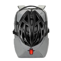A gray bicycle backpack shown from the front, featuring a black bicycle helmet mounted on the back and a small red rear light. The backpack has gray webbing straps and a top handle, photographed against a solid white background.