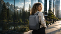 A woman with curly hair stands on a city sidewalk next to a reflective glass wall showing a mountain and forest scene. She is wearing a light gray backpack, which is the product being showcased in this lifestyle setting.