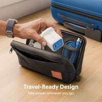 A hand inserts a white and blue portable charger into a dark gray zippered travel pouch on a light wood table, with a blue suitcase in the background and the text 'Travel-Ready Design' at the bottom.