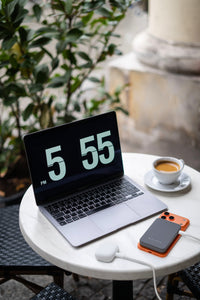 A silver laptop sits on a round white table outdoors, displaying 5:55 on its screen. A cup of coffee, a white earbud, and an orange-and-gray phone case are also on the table.